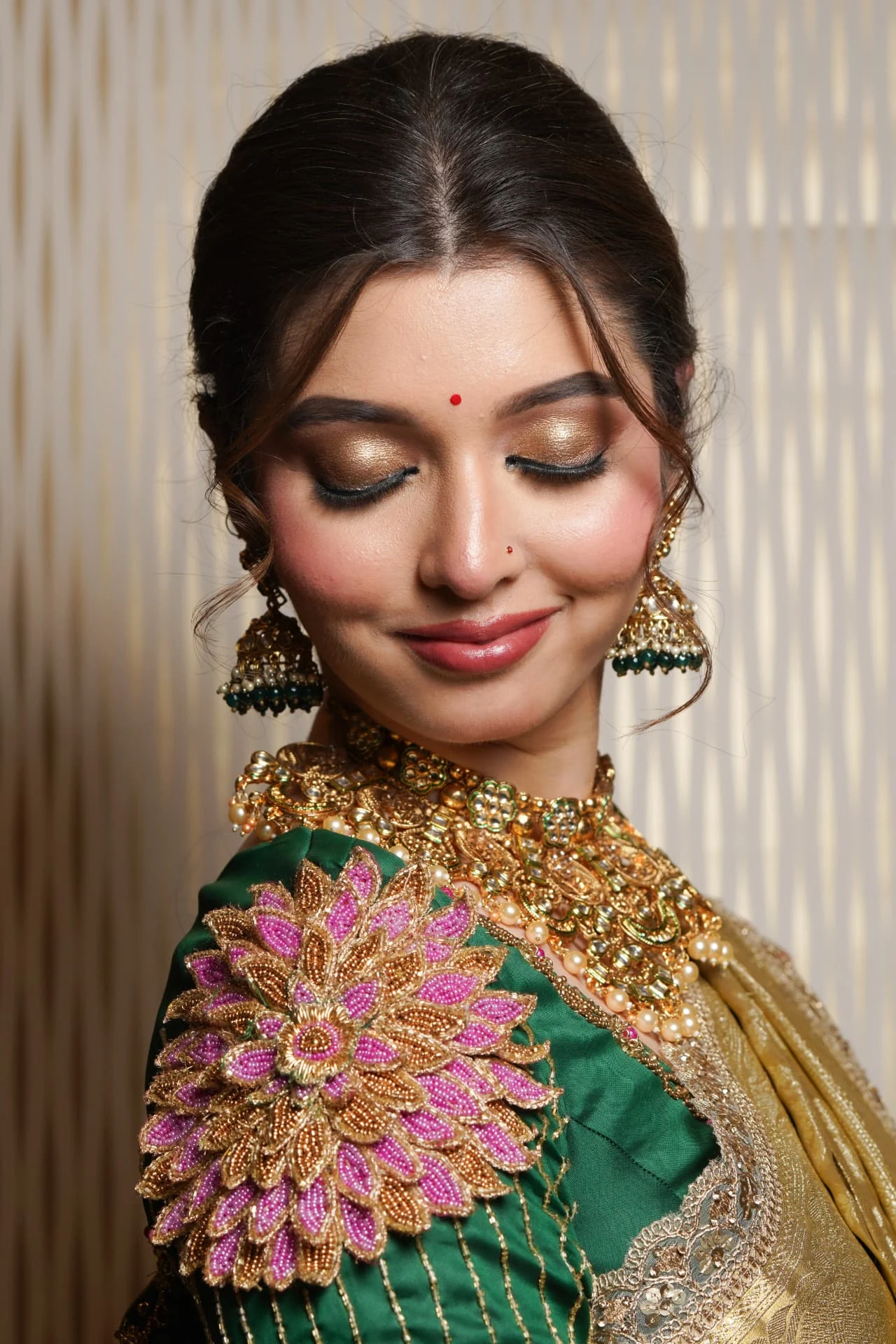 Bride with eyes closed in an emerald green bridal blouse with pink beaded shoulder embroidery, gold kundan choker and jhumka earrings.